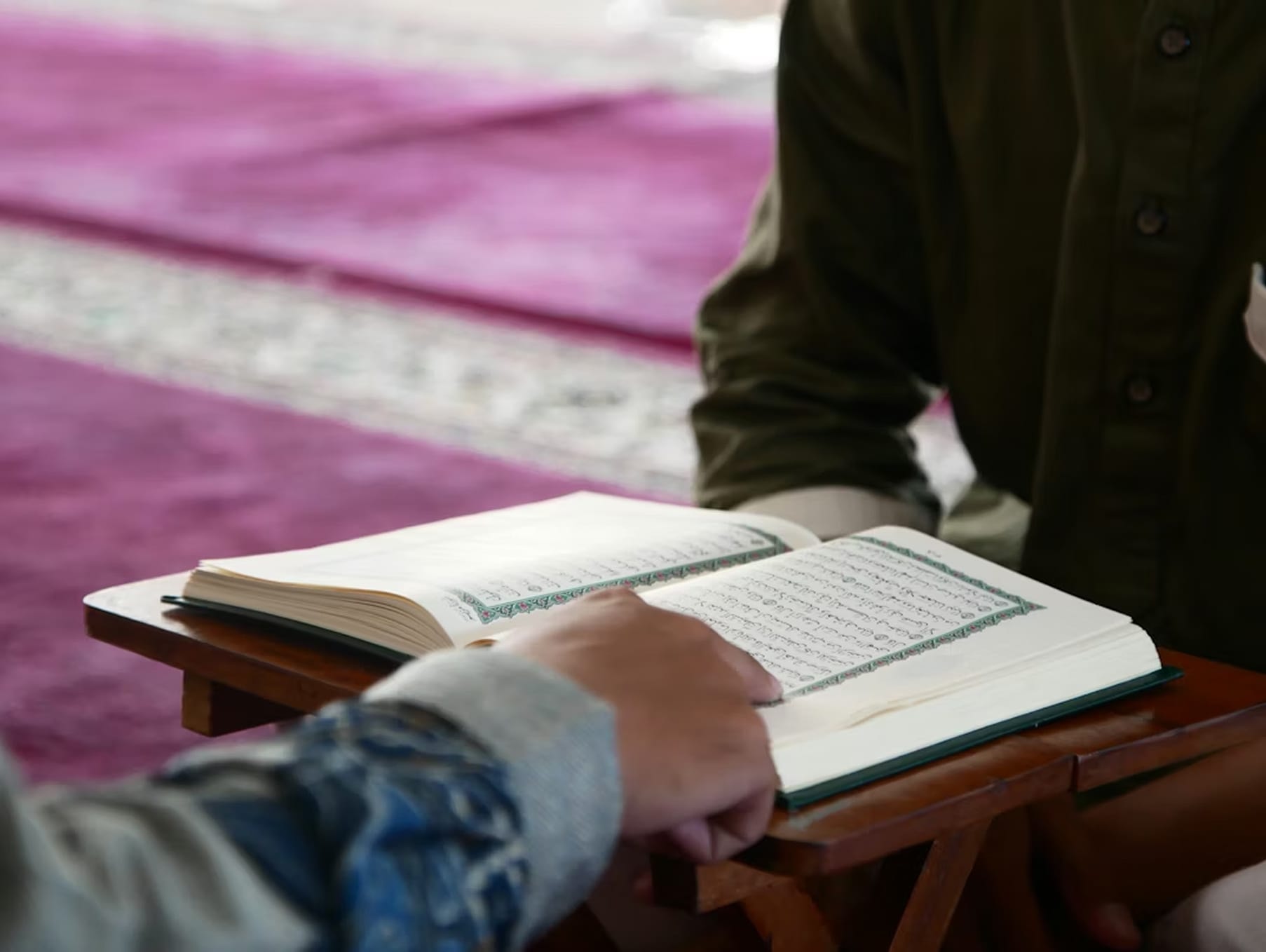 Hands resting on an open Qur'an at a wooden stand in the prayer hall